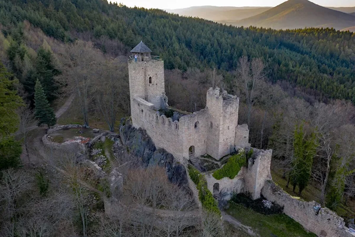 Vue aérienne du château du Bernstein : donjon en granite coiffé d’un belvédère, logis et enceinte en forêt au-dessus de Dambach-la-Ville