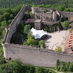 Vue aérienne du château du Hohlandsbourg : grande enceinte et chemin de ronde, cour sous auvent blanc et château supérieur sur son rocher, en forêt