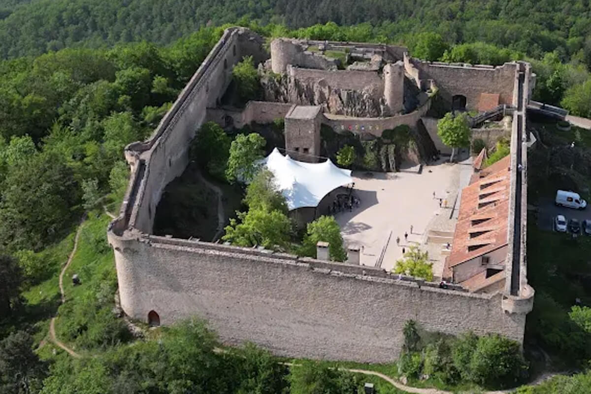Vue aérienne du château du Hohlandsbourg : grande enceinte et chemin de ronde, cour sous auvent blanc et château supérieur sur son rocher, en forêt