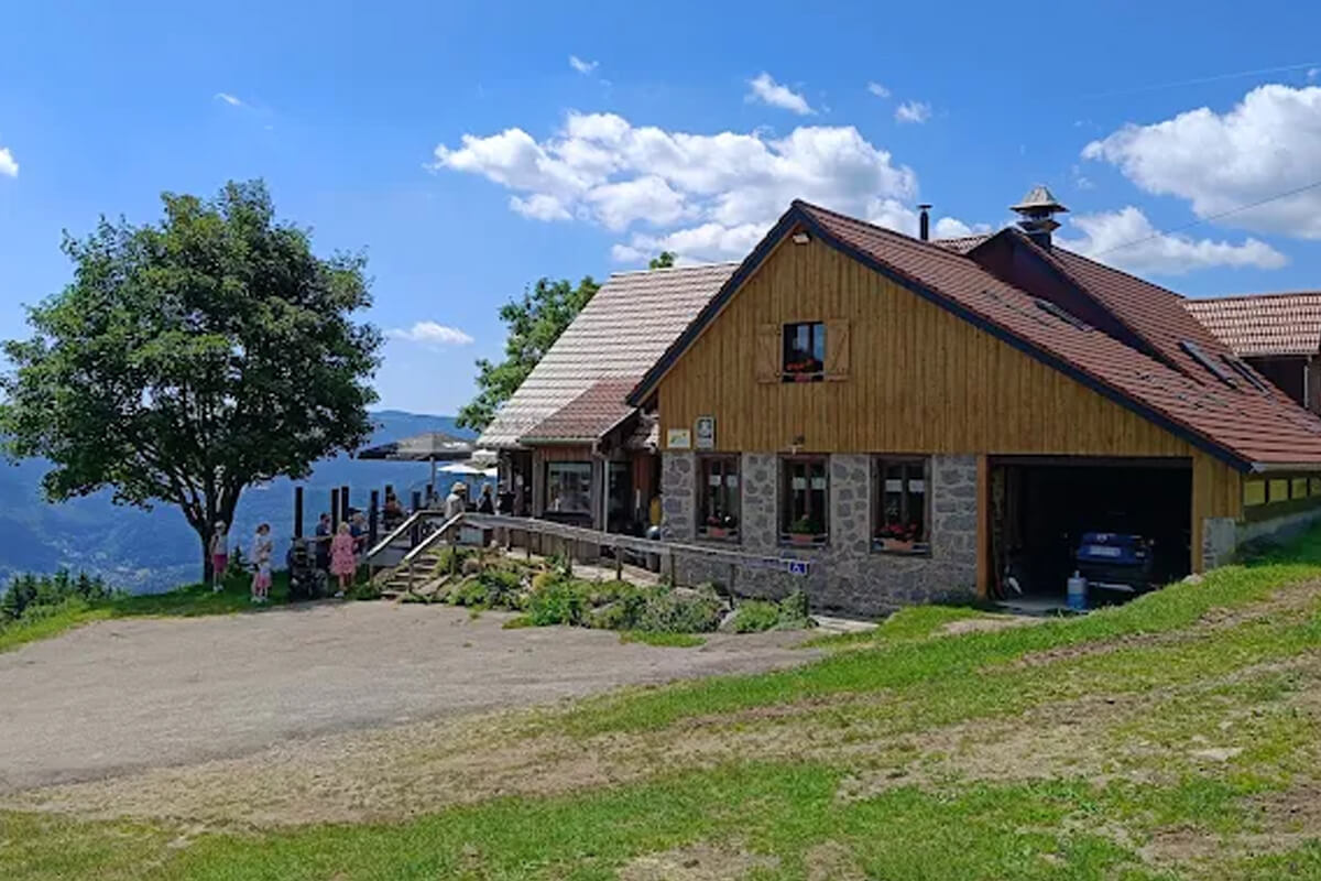 Ferme-auberge de montagne au bardage de bois clair sur soubassement de pierre, terrasse animée et vue sur une vallée des Vosges en été